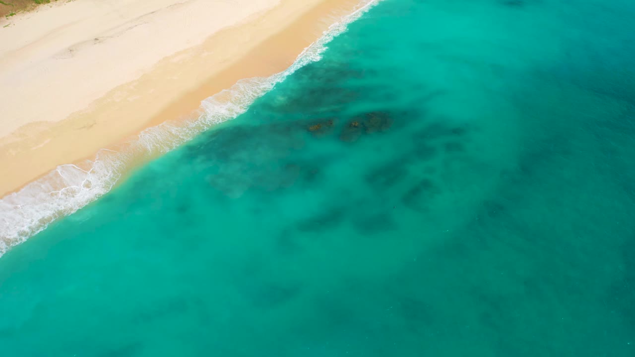 Aerial frame in Baja California Sur with the sandy shoreline running diagonally, gentle white wash at the waterline, dark shapes under the clear water, and no visible people.