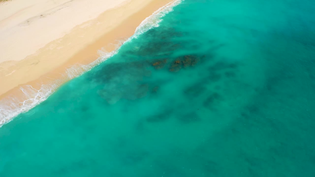 High-angle drone view in Baja California Sur where pale beach curves along the top left, blue-green water dominates the frame, darker underwater formations sit offshore, and no visible people appear.