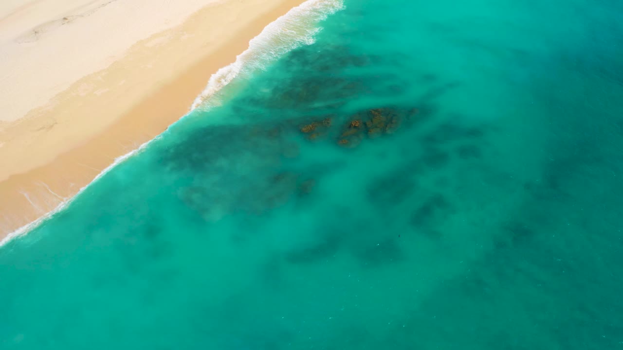 Overhead Baja California Sur coastline with sand along the left edge, white surf curling into shallow turquoise water, larger dark submerged patches near center, and no visible people.