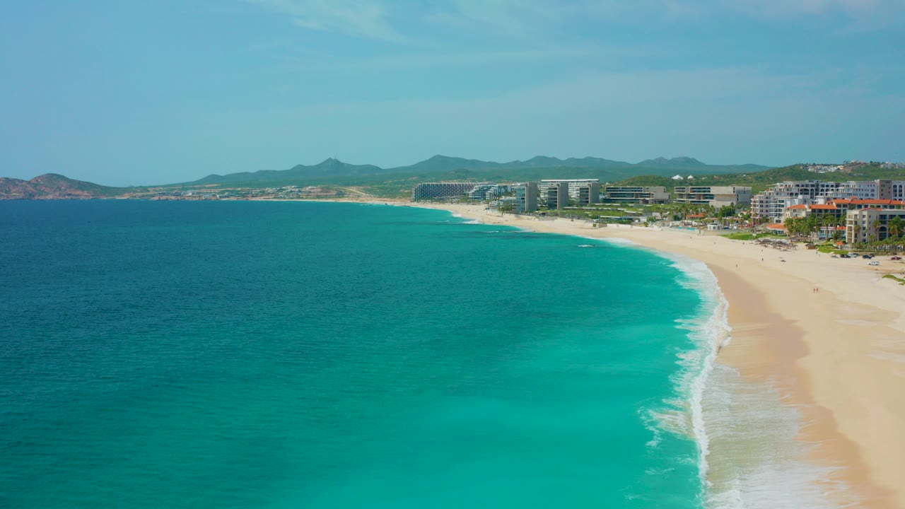 High-angle aerial frame over Desert Park Natural Reserve in Baja California Sur, with turquoise water on the left, a pale sandy beach on the right, beachfront buildings beyond the sand, distant hills, and no visible people.