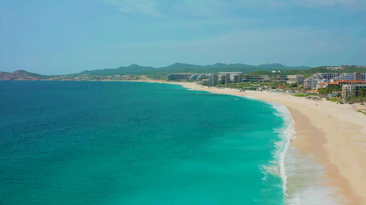 Aerial view in Baja California Sur showing the surf line curving along the sand, deep blue water at left, lighter turquoise shallows near shore, modern beachfront buildings in the distance, and no visible people.