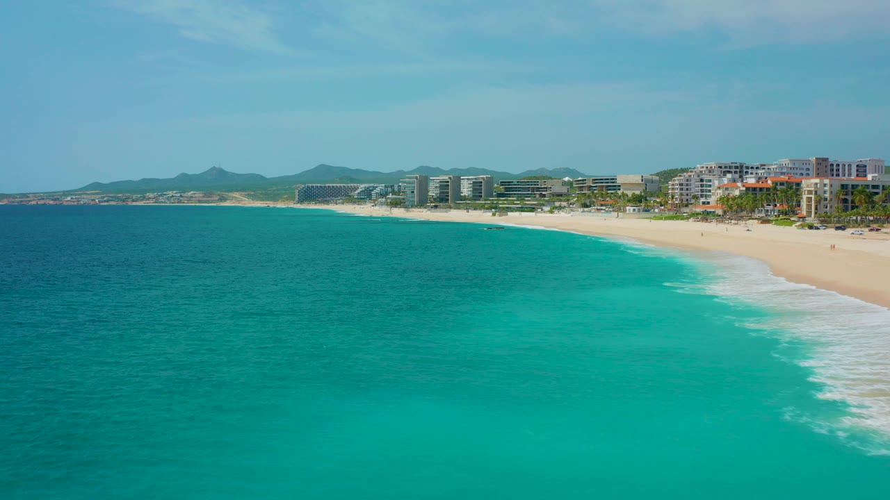 High aerial frame from Desert Park Natural Reserve, Baja California Sur, Mexico, with the turquoise sea dominating the foreground, surf tracing the beach, distant hills, and no visible people.