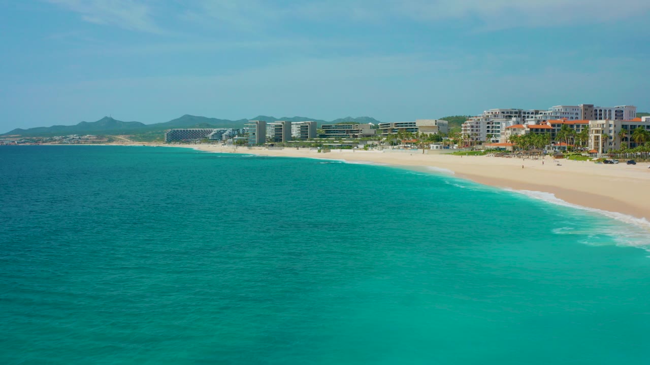 Opening aerial frame over Desert Park Natural Reserve, Baja California Sur, with turquoise ocean in front, a pale beach and hotel row beyond, distant hills, clear sky, and no visible people.