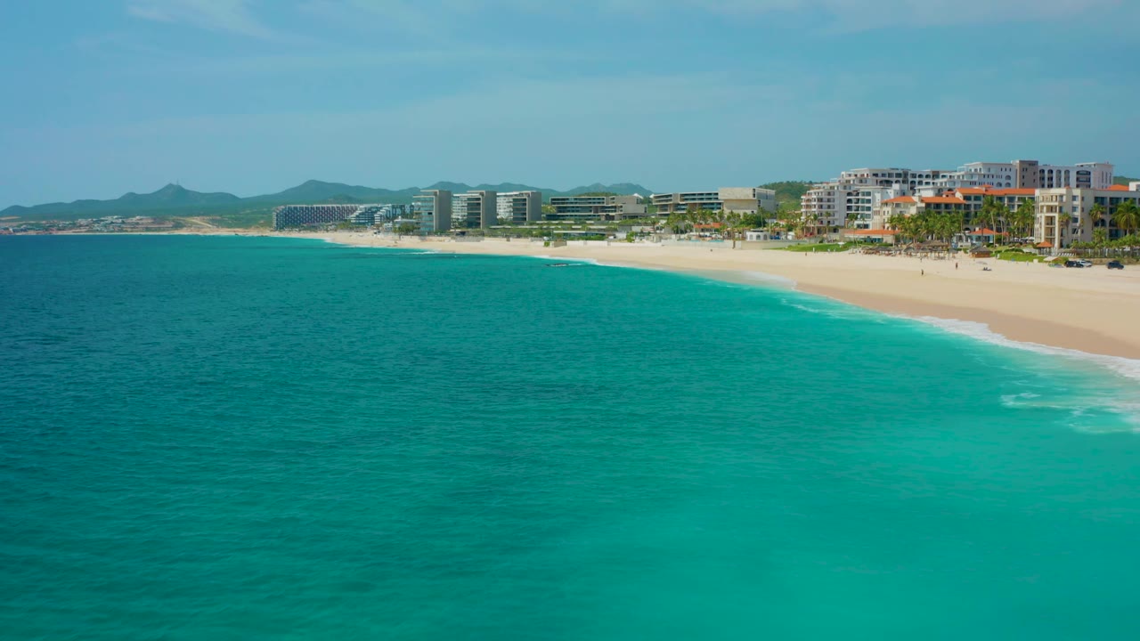 Aerial frame at 0:02 over Desert Park Natural Reserve, Baja California Sur, with the turquoise sea dominating the foreground and the sandy resort beach and hotel buildings pushed toward the upper edge, with no visible people.