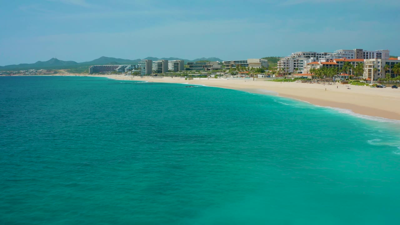 Aerial resort shoreline with turquoise water, pale sand, hotels, and hills.