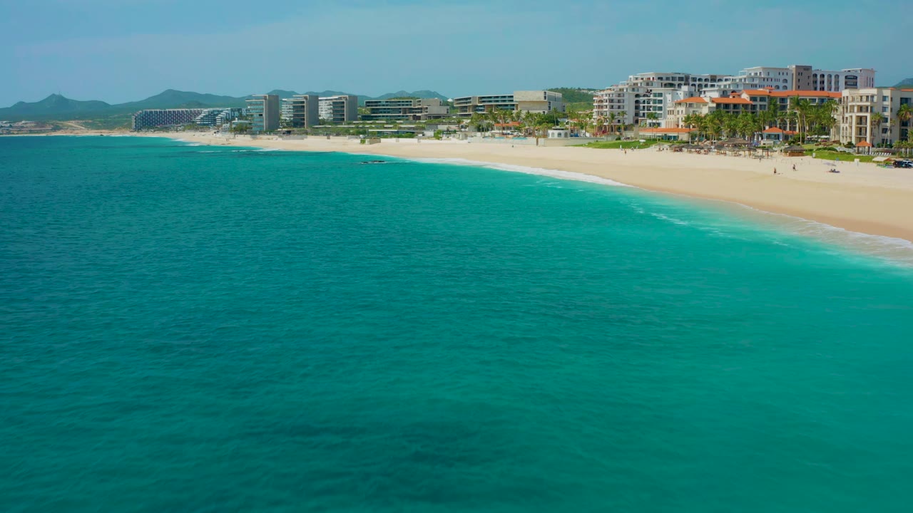 Aerial frame one second in with calm turquoise water below a curved sandy beach and multi-story resort buildings in Desert Park Natural Reserve, Baja California Sur, Mexico, with no visible people.
