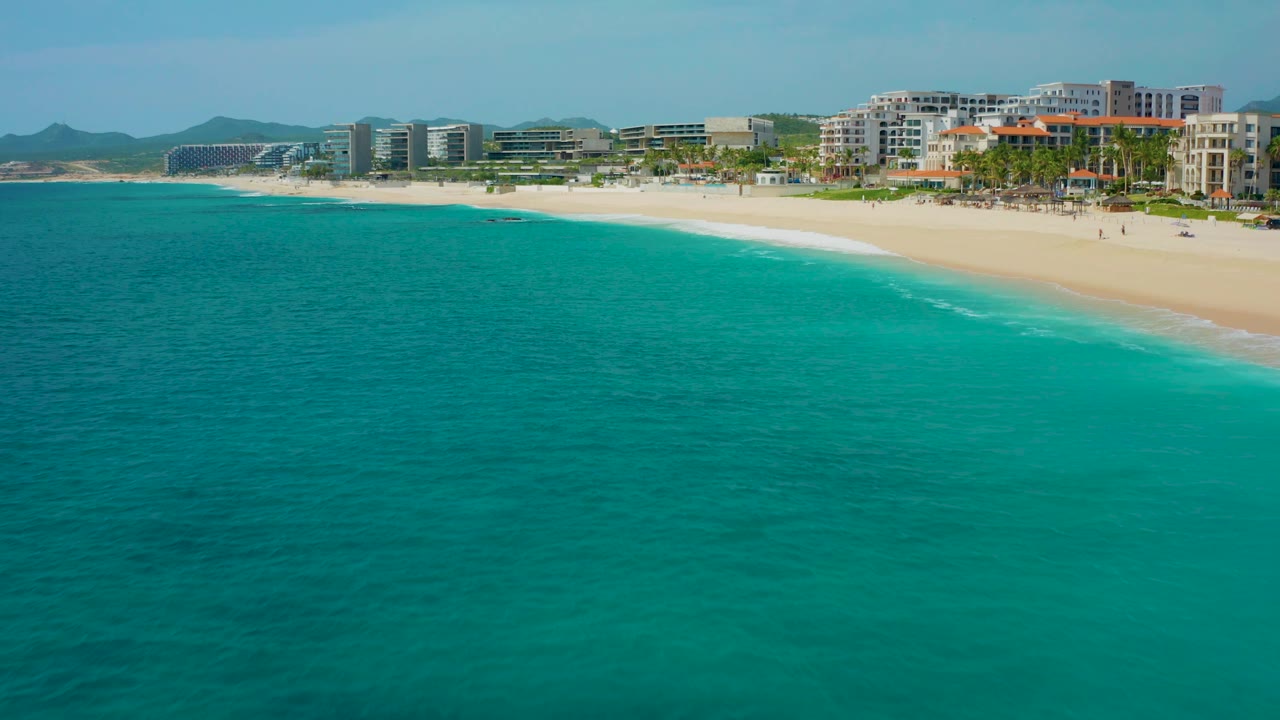 Drone-height view across teal ocean toward a bright sandy beach and resort-lined shore in Desert Park Natural Reserve, Baja California Sur, Mexico, with no visible people.
