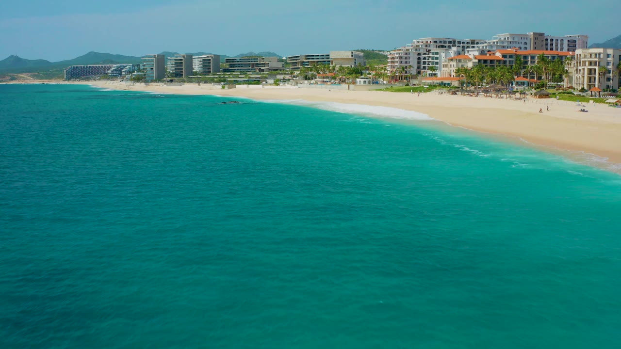 Mid-clip aerial view of the beach curve, turquoise water, pale sand, and shoreline resorts in Desert Park Natural Reserve, Baja California Sur, Mexico, with no visible people.