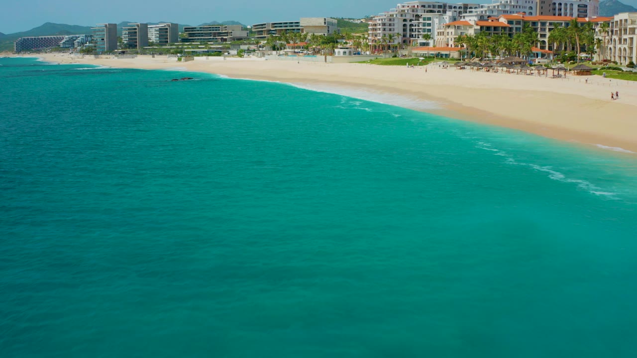 Aerial frame from Desert Park Natural Reserve in Baja California Sur showing turquoise ocean in the foreground, pale beach across the top, hotel-like buildings, palms, distant hills, and no visible people.