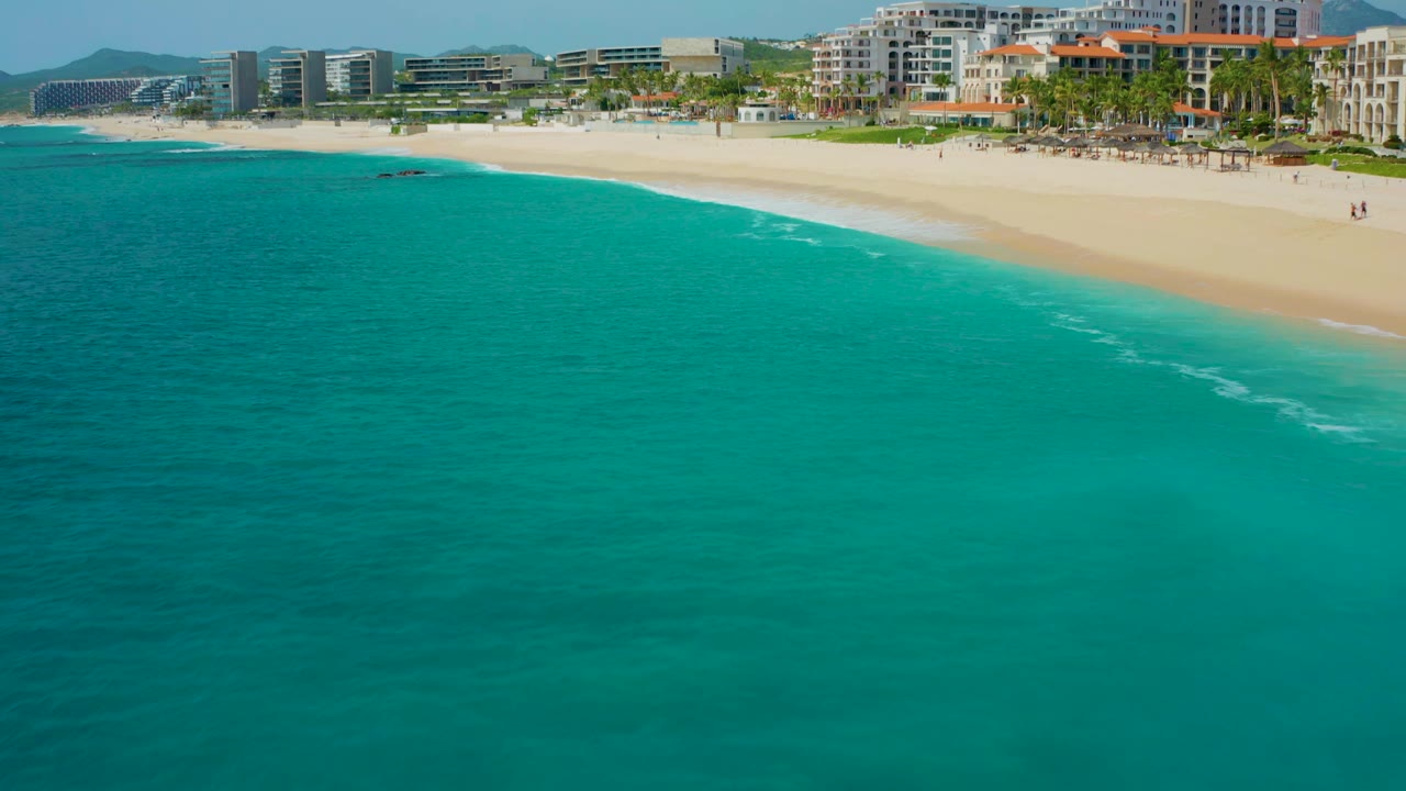Drone view over turquoise water toward a bright sandy beach with buildings behind it.