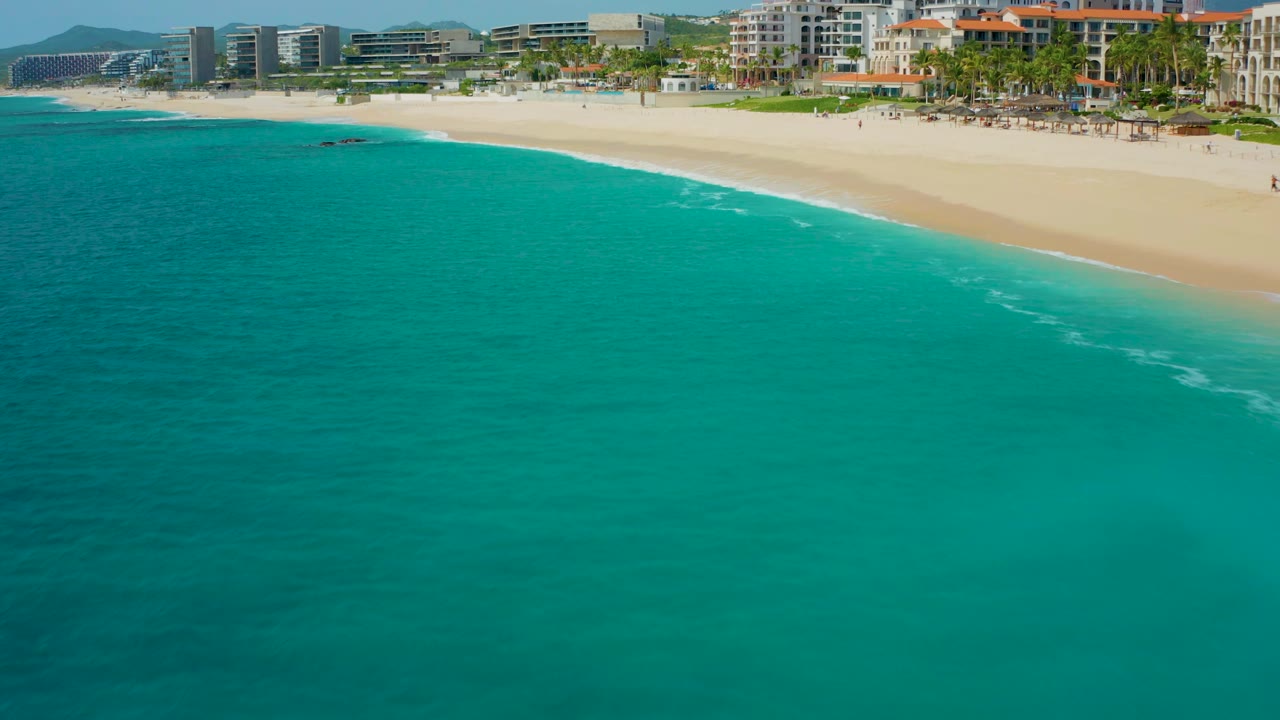 Wide aerial frame from Baja California Sur with teal ocean covering the lower field, pale beach above it, a few white wave marks, palm-lined development, distant mountains, and no visible people.