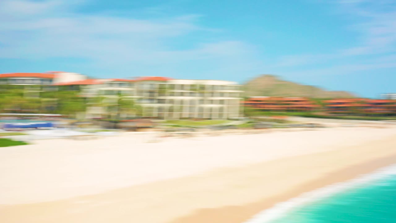 Opening aerial frame over Desert Park Natural Reserve in Baja California Sur, with motion-blurred resort roofs, pale beach, turquoise water, and no visible people.