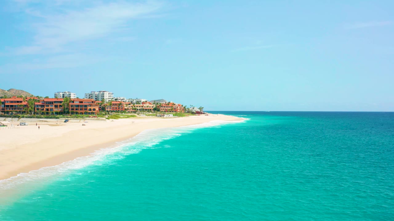 Mid-clip aerial frame over Desert Park Natural Reserve in Baja California Sur with resort buildings set back from the beach, broad turquoise water in front, and no visible people.