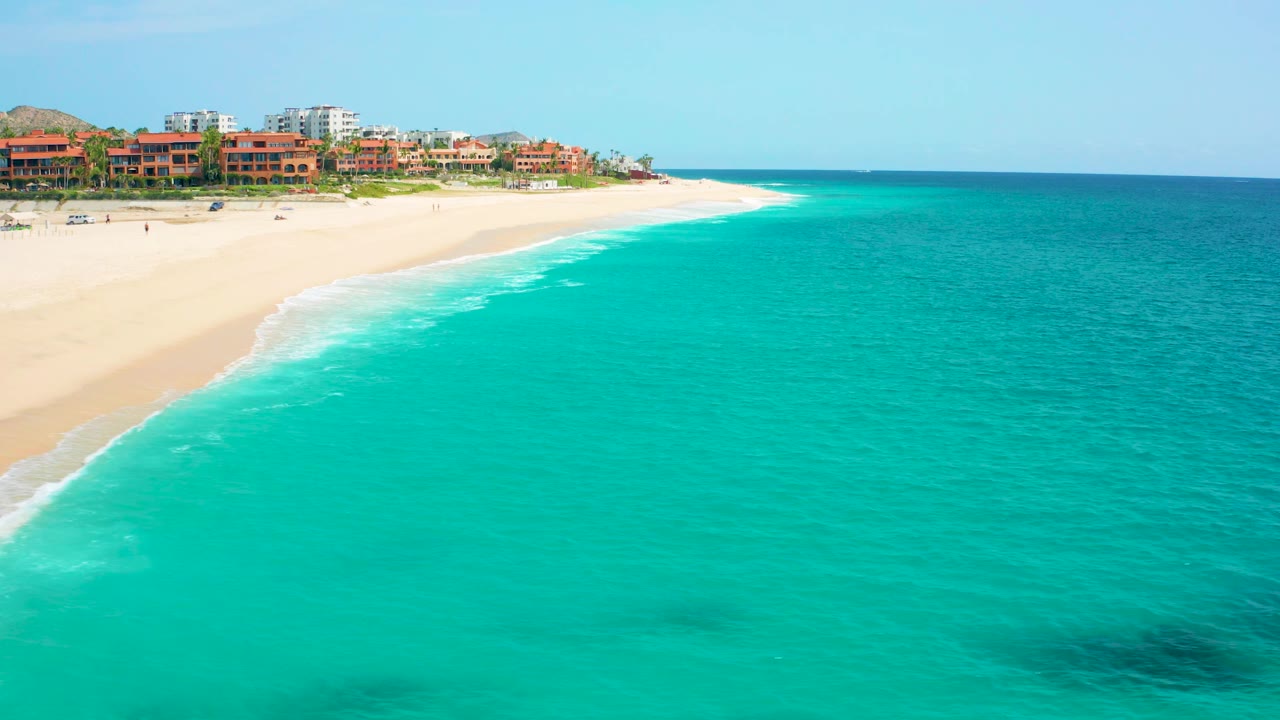 High-angle aerial frame over Desert Park Natural Reserve in Baja California Sur, Mexico, showing turquoise ocean, pale sand, white surf, beachfront buildings, and no visible people.