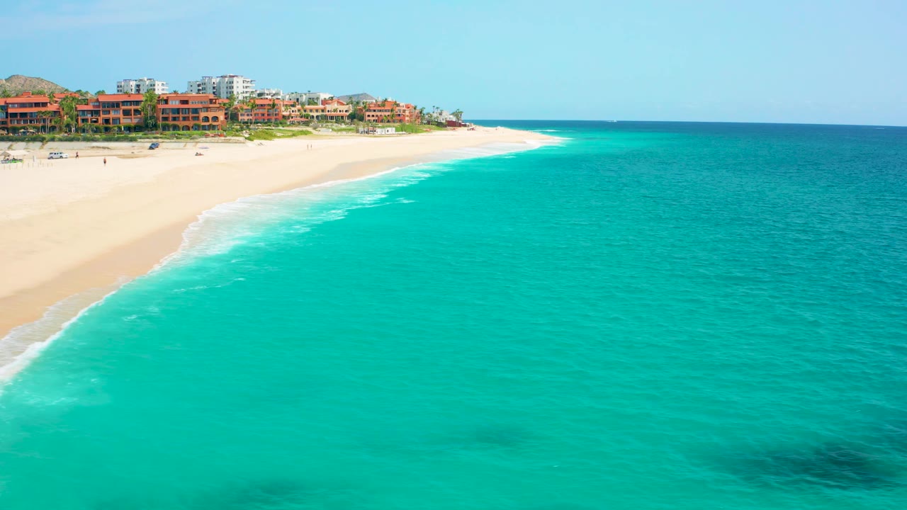 Aerial view in Baja California Sur, Mexico, with the shoreline curving from lower left toward distant beachfront buildings and no visible people on the sand.