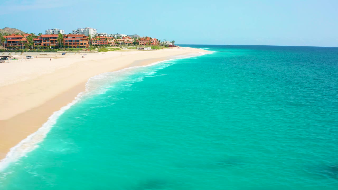 High aerial frame from Desert Park Natural Reserve, Baja California Sur, Mexico, showing open beach, blue-green ocean, low surf, distant buildings, and no visible people.