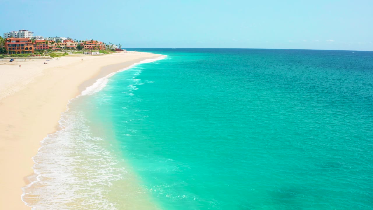 Aerial frame from Baja California Sur showing turquoise water, pale sand, white surf, clear sky, and distant resort-style buildings along the shoreline with no visible people.