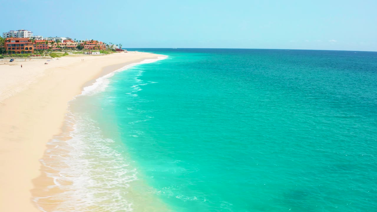Wide aerial view in Baja California Sur with open turquoise sea filling the right side, pale sand and breaking surf on the left, and terracotta-roof buildings near the upper shore with no visible people.