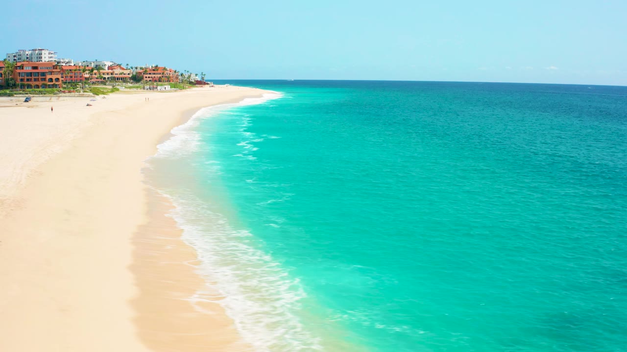 Baja California Sur aerial showing white foam running diagonally along the sand, bright turquoise water to the right, distant beachfront buildings above the beach, and no visible people.