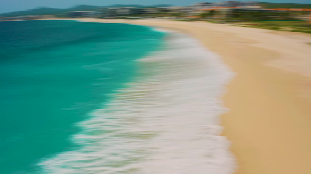 Early aerial frame over the Desert Park Natural Reserve coastline in Baja California Sur, with motion-blurred turquoise water, pale sand, white surf, and no visible people.