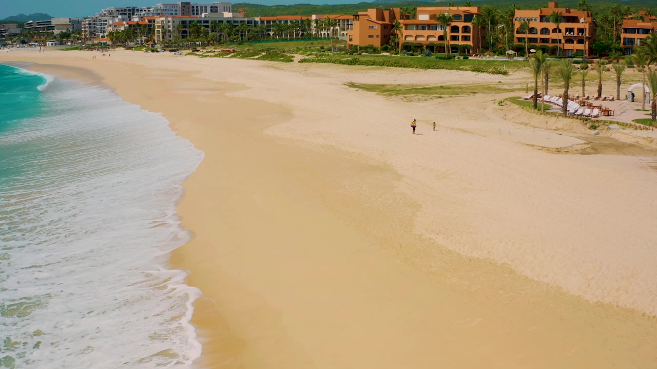 Aerial frame at Desert Park Natural Reserve, Baja California Sur showing pale sand, shallow surf, palm-lined coastal buildings, low hills, and no clearly visible people.