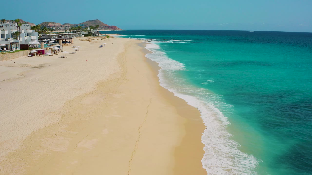 Opening aerial frame over Desert Park Natural Reserve in Baja California Sur, with wide tan sand at left, turquoise ocean at right, white surf along the shore, beachfront buildings at the far left, and no visible people.