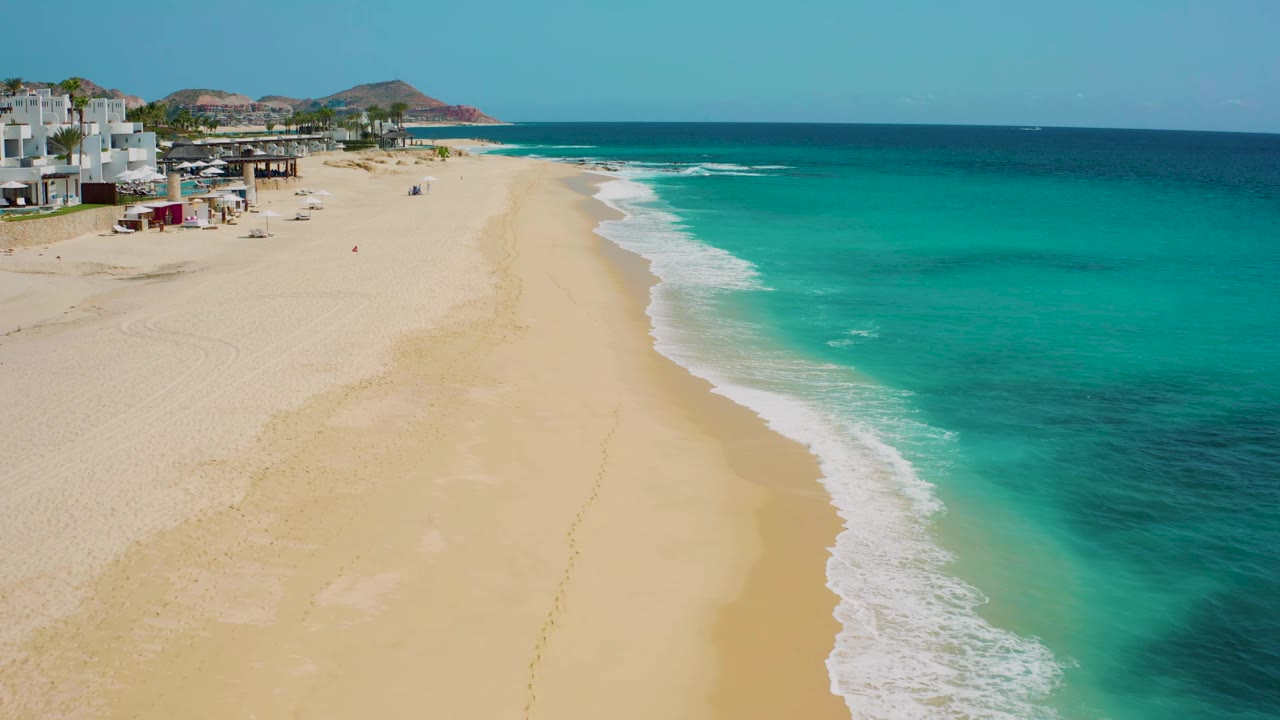 Aerial view along the Baja California Sur shoreline, with the beach stretching into the distance, surf forming a curved white edge, low headlands on the horizon, and no visible people.