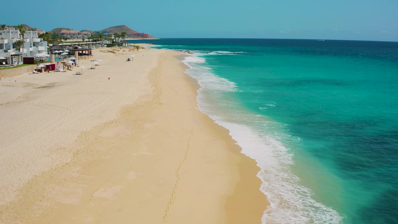 The drone view holds the Desert Park Natural Reserve beach from above, showing white buildings and small beach structures at left, a broad empty sand strip, turquoise water, and no visible people.