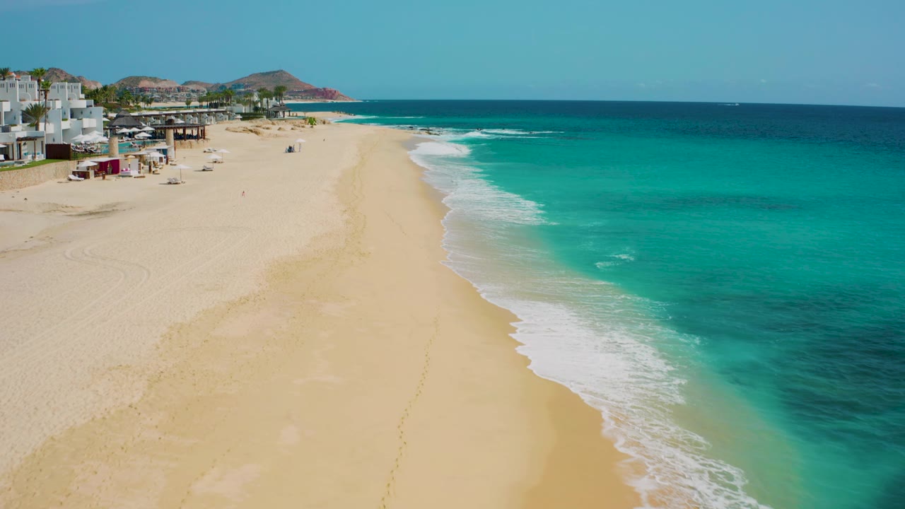 Aerial coastline frame with tan sand, turquoise sea, and white surf.