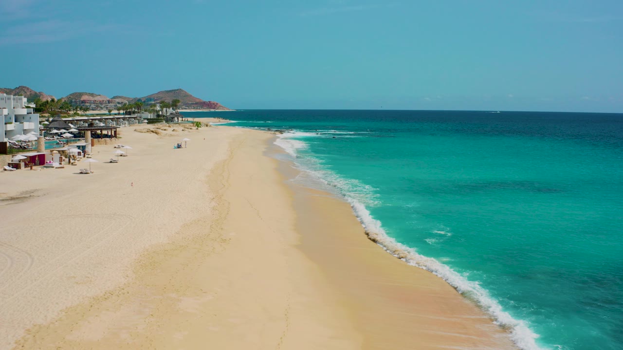 High aerial frame of Desert Park Natural Reserve in Baja California Sur, with pale sand curving beside turquoise water, beachfront buildings at the far left, low hills on the horizon, and no visible people.