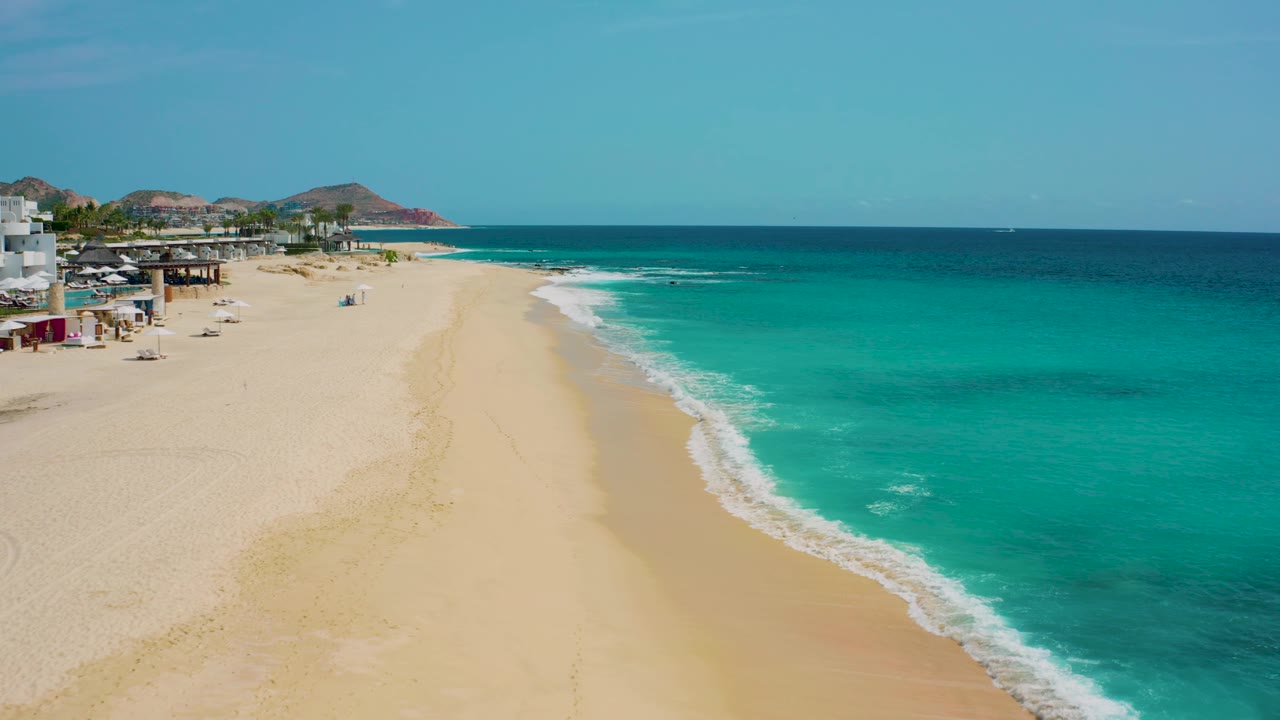 The coast at Desert Park Natural Reserve remains framed from above, with beachfront structures and palms at upper left, a wide empty sand strip, bright aqua water, and no visible people.