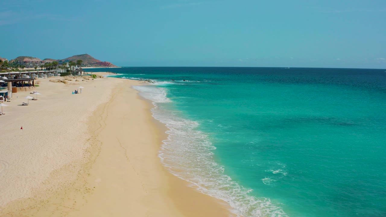 Aerial frame over Desert Park Natural Reserve in Baja California Sur, Mexico, showing a broad sandy beach, turquoise shore water, deeper blue ocean, distant rocky hills, and no visible people.