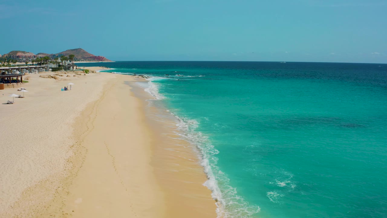 Aerial view in Desert Park Natural Reserve, Baja California Sur, Mexico, looking along empty-looking sand with a bright white surf line, aquamarine shallows, darker sea beyond, and no visible people.