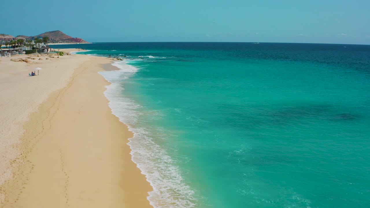 Aerial frame from Desert Park Natural Reserve in Baja California Sur showing turquoise water, white foam, a broad sandy beach, distant hillside, and no visible people.