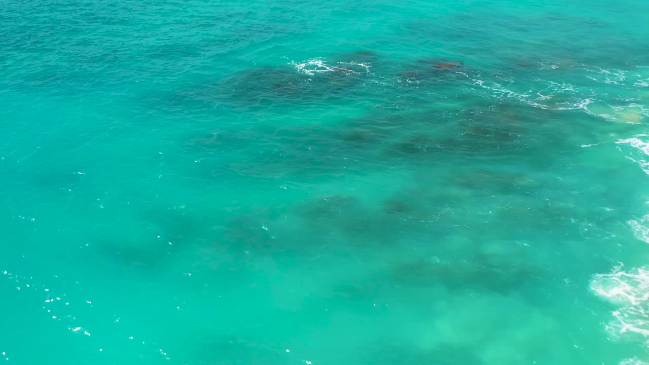 Aerial view of clear turquoise water in Baja California Sur, showing darker underwater formations spread across the upper half and no visible people.