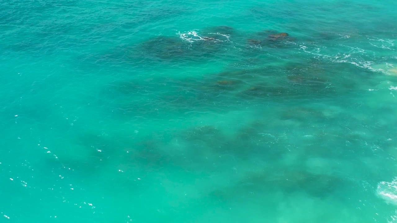 Overhead drone frame of bright aqua ocean at Desert Park Natural Reserve, with reef-like dark patches visible through shallow water and no visible people.