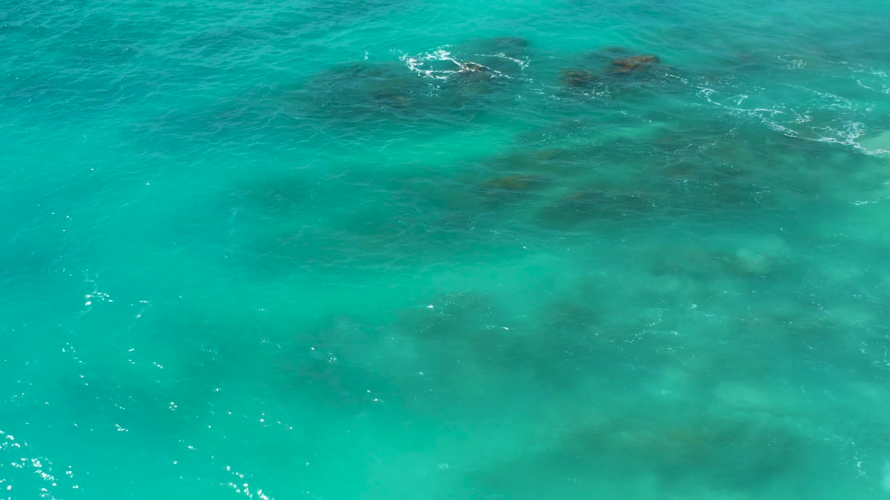High-angle aerial still over turquoise sea in Baja California Sur, with scattered sun glints, darker submerged patches, and no visible people or boats.
