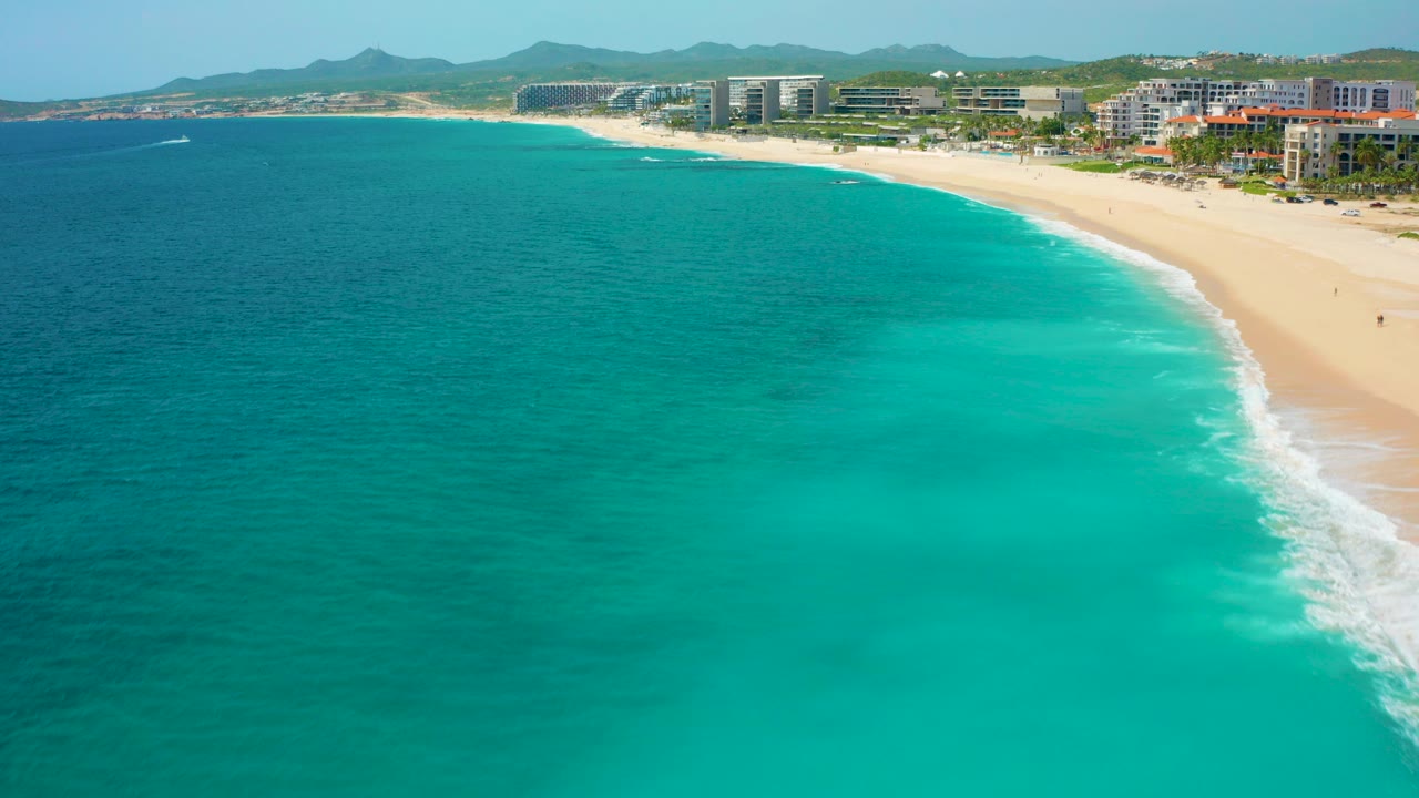 Opening aerial frame over Desert Park Natural Reserve in Baja California Sur, Mexico, showing turquoise water, a curving sandy beach, white surf, beachfront buildings, distant hills, and no visible people.