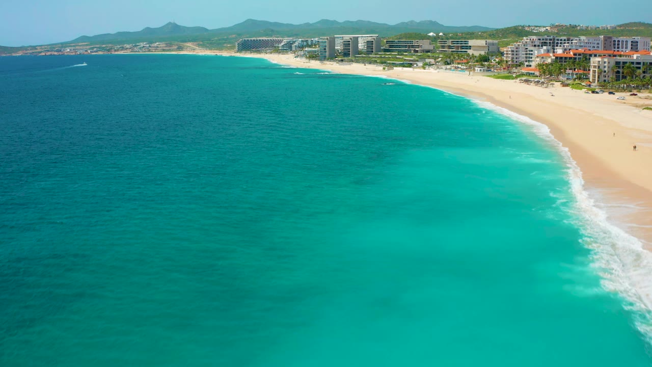 Aerial view in Baja California Sur, Mexico, with blue-green water covering the foreground, a pale beach along the right edge, hotel-like buildings near the shore, distant hills, and no visible people.