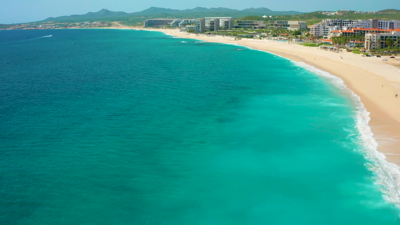 High aerial frame over Desert Park Natural Reserve showing darker offshore water shifting into turquoise shallows, a narrow surf line, sandy beach, modern coastal buildings, hills, and no visible people.