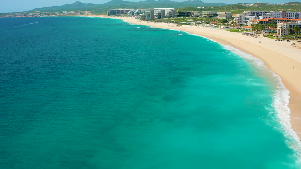 Mid-clip aerial view in Baja California Sur, Mexico, with open ocean across the left side, turquoise water near shore, a curved sandy beach to the right, beachfront buildings, hills, and no visible people.