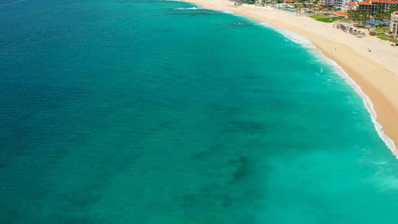 High drone frame of Desert Park Natural Reserve coastline, with the beach curving across the upper right, clear turquoise water below, and resort-style buildings at the shore with no visible people.
