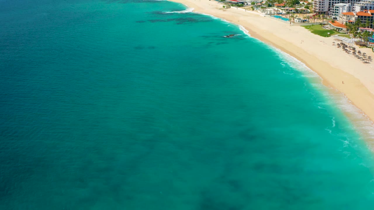 Aerial frame from Baja California Sur showing broad turquoise water, a diagonal sandy shoreline, beachfront buildings, and no visible people.