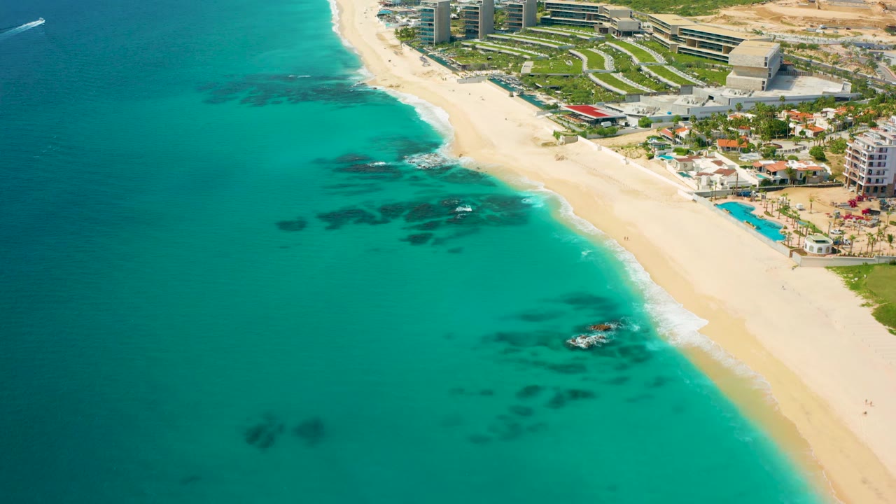 High-angle coastal frame in Baja California Sur showing clear turquoise water, pale sand, dark reef-like patches offshore, hotel buildings, and no visible people.