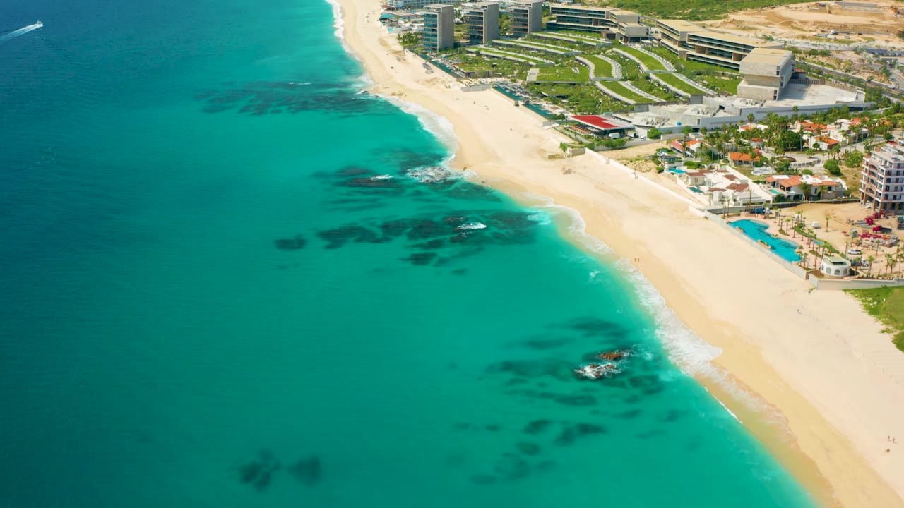 Aerial frame over the Baja California Sur shoreline with the beach running diagonally, dark underwater rock patches offshore, resort grounds above the sand, and no visible people.