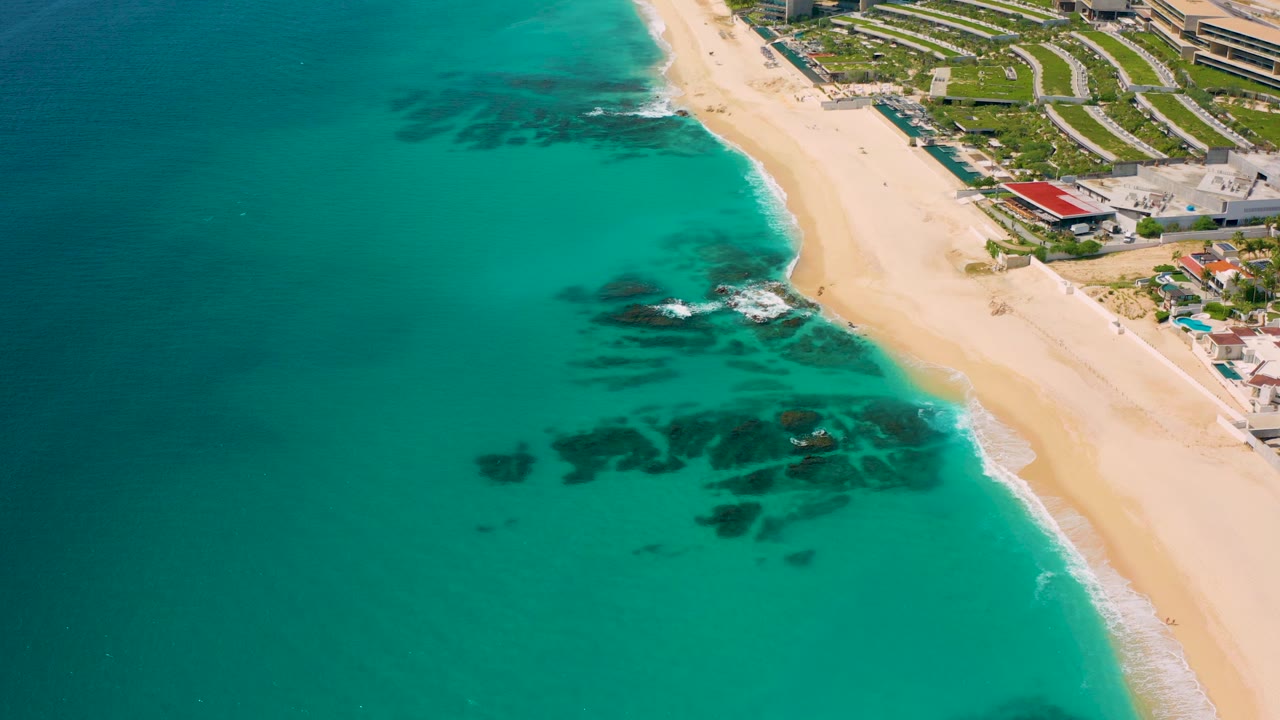 Aerial view at Desert Park Natural Reserve in Baja California Sur, Mexico, showing turquoise water, pale sand, dark underwater rocks, resort-style buildings, and no visible people.