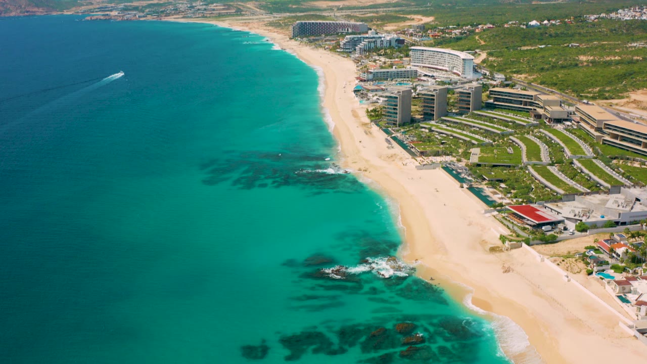 High-angle aerial view in Desert Park Natural Reserve, Baja California Sur, showing turquoise water beside a wide sandy beach, resort buildings onshore, and a small boat wake offshore, with no visible people.