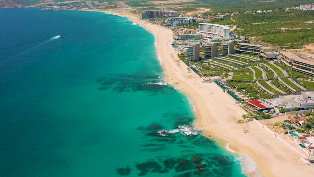 Aerial frame over Desert Park Natural Reserve, Baja California Sur, with the beach running diagonally between clear turquoise ocean and resort buildings, while the boat wake remains left of shore and no visible people appear.