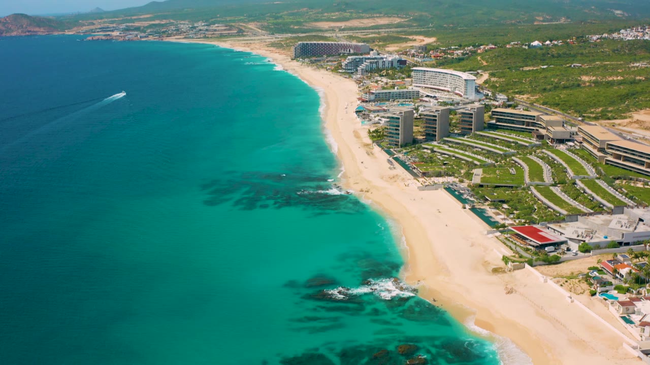 High aerial view in Desert Park Natural Reserve, Baja California Sur, showing the resort district along the sand, darker reef-like patches in turquoise water, and inland hills beyond the green terrain, with no visible people.