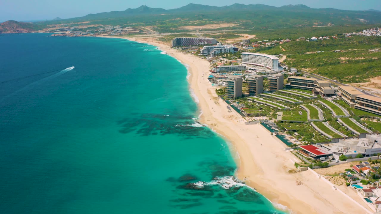 Wider aerial frame over Desert Park Natural Reserve, Baja California Sur, with more inland hills visible above the resort buildings and a small white wake crossing the turquoise water, with no visible people.